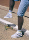 A person wearing M0007 by Adriano and light blue socks, lifting one foot onto a curb, dressed in light blue denim shorts on a brick pavement with a background of brown bricks.