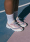 A person wearing white socks and pastel-colored L0018 sneakers by Bruna, handcrafted from premium Italian leathers, stands on a multi-colored sports court.