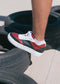 A person wearing N0016 by Daniel handcrafted sneakers in red, black, and white stands on a stacked pile of tires on a tiled surface, showcasing the premium Italian leathers used in their ethically made footwear.