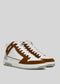 The V6 Caramel W/ White is a pair of high-top sneakers meticulously handcrafted in Portugal. They are made from premium Italian leathers, showcasing a stylish combination of brown suede and white leather panels. Equipped with white laces and rubber soles, these sneakers are artfully positioned at an angle on a plain background.