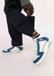 Person sporting V1 Petrol Blue w/ White sneakers handcrafted in Portugal, paired with striped socks and dark pants, adjusting their pant leg.