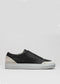 The V19 Black Leather W/ Bone is a slip-on sneaker with a premium leather upper, combining black and white hues, showcased against a plain gray background. It features a pristine white rubber sole.