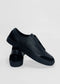 A pair of SO0025 Black Leather slip-on sneakers, handcrafted in Portugal from premium Italian leathers, with one shoe standing and the other leaning against it on a white background.