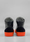 Rear view of the MH0121 Grey Floater, a pair of high-top shoes featuring black leather uppers, black suede detail on the heel, and vibrant orange rubber soles against a plain gray background. Handcrafted in Portugal using premium Italian leathers, these shoes are ethically made to order for a unique touch.