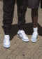 Two people standing on a sidewalk wearing light blue M0011 by Serifo and white socks. One person is sporting black cargo pants while the other has on dark gray shorts. These stylish kicks, handcrafted in Portugal, effortlessly elevate their casual streetwear look.