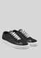 The V1 Black sneakers are low-top shoes with white soles and laces, crafted from premium Italian leathers. Expertly handcrafted in Portugal, these exquisite shoes are elegantly positioned on a gray background.