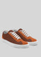 The V7 Brown & Caramel, a pair of low-top sneakers crafted from premium Italian leathers in orange and brown, with white laces and soles, is placed on a light gray background.