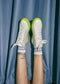 Legs raised against a blue curtain, wearing MH0088 White Leather W/ Lime with neon green soles and purple striped socks. Lower legs have tattoos. The handcrafted sneakers boast premium Italian leathers, offering both style and sustainability for the ethically-minded individual.