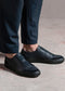 A person is wearing SO0025 Black Leather made from premium Italian leathers with black pants. The shoes, handcrafted in Portugal, feature a textured surface and elastic sides for ease of wear. The backdrop is a concrete floor.