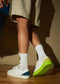 A person wearing ML0038 White Leather sneakers with premium Italian leathers that feature different colored backs in blue and yellow, along with neon green soles, paired with long white socks and beige shorts, walking on a wooden floor.