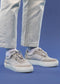 A person wearing N0024 White sneakers handcrafted in Portugal and white socks stands on a blue background, donning light-colored cuffed pants.